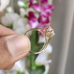 a person holding a gold ring with a flower in the middle and pink flowers in the background