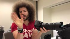 a man with an afro is holding a camera in front of him and making the peace sign