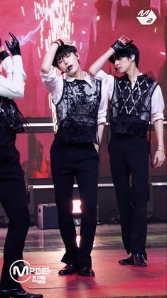 three men in black pants and white shirts are standing on stage with their hands behind their head