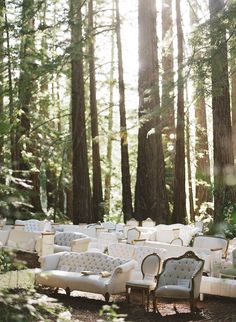 an outdoor seating area in the woods with white couches and chairs set up for a wedding