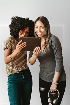 two young women standing next to each other holding an electronic device and smiling at the camera