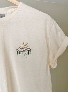 a white t - shirt with trees and mountains embroidered on the front, hanging from a wooden hanger