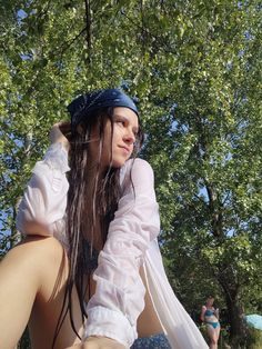 a woman with long hair sitting on the ground in front of trees and wearing a white shirt