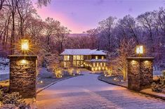 a large house surrounded by trees covered in snow at night with lights on the driveway