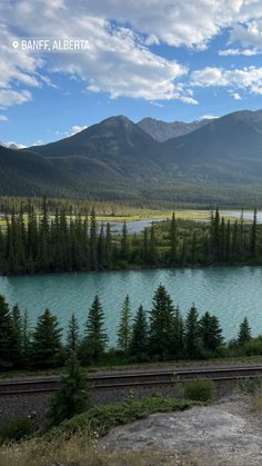 there is a train that is going by the water and mountains in the distance with trees on both sides