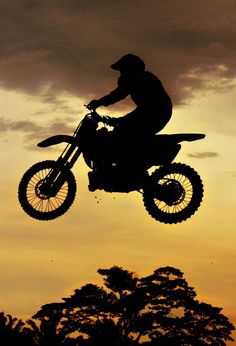 a person on a dirt bike jumping in the air at sunset or dawn with trees and clouds behind them