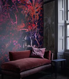 a red couch sitting in front of a window next to a wall covered in flowers