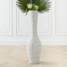 a white vase filled with green plants on top of a hard wood floor next to a wall