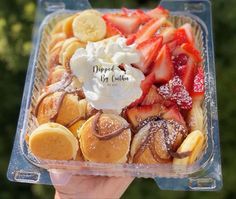 a person holding up a dessert in a plastic container with strawberries, bananas and whipped cream on top