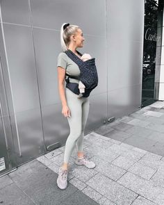 a woman is standing on the sidewalk with her baby in a sling carrier and looking at something