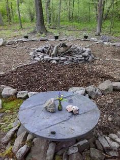 a table with rocks and flowers on it in the middle of a wooded area, surrounded by trees