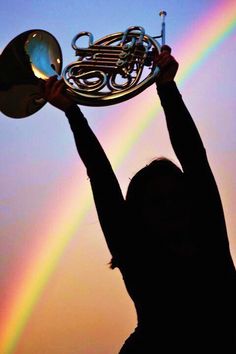 a woman holding up a french horn with a rainbow in the background