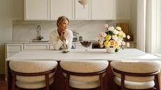 a woman sitting at a kitchen table with four stools in front of her and flowers on the counter