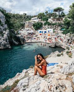 a woman sitting on top of a rocky cliff next to the ocean taking a photo