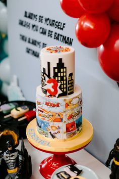 a three tiered cake decorated with comic characters and balloons in the shape of batman