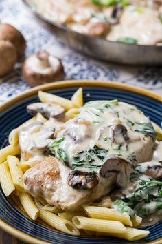 a blue plate topped with pasta covered in mushrooms and sauce next to a pan of chicken