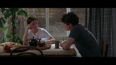 two people sitting at a table talking to each other in front of a potted plant