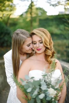 two beautiful young women hugging each other in front of some greenery and trees at the end of their wedding day