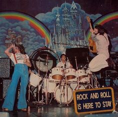 the rock and roll band is performing in front of a stage backdrop that says rock and roll is here to stay