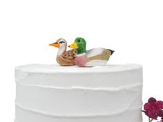 two ducks sitting on top of a white cake next to a purple flower in front of a white background