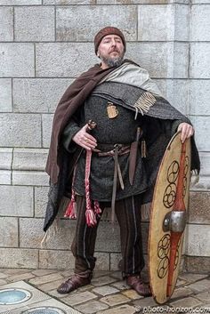 a man dressed in medieval clothing standing next to a stone wall with a shield on it