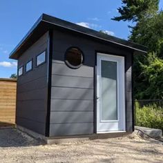 a small gray shed with a white door and window on gravel ground next to trees