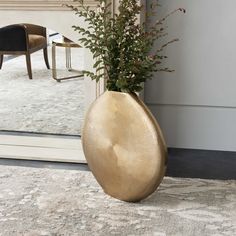 a plant in a gold vase sitting on a rug next to a chair and mirror