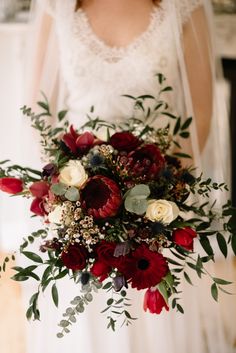 a bride holding a bouquet of red and white flowers with greenery in her hand