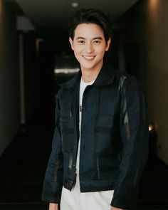 a young man standing in a hallway wearing a black jacket and white pants with his hands in his pockets