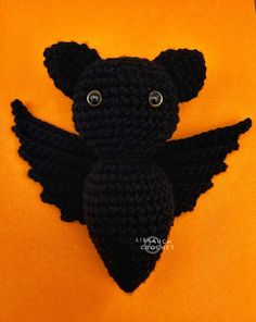 a crocheted black teddy bear with wings on an orange background, looking up at the camera