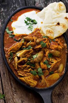 some food is in a black bowl on a wooden table next to bread and sour cream