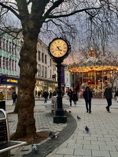 a clock on a pole in the middle of a street with people walking around it