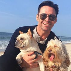 a man sitting on the beach with two dogs in his lap and smiling at the camera