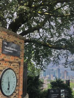 a clock on the side of a brick wall under a tree in front of a cityscape