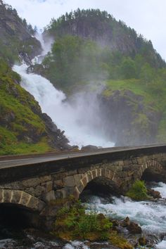 a stone bridge over a river next to a lush green hillside with waterfall in the background