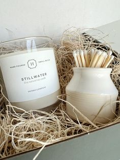 a candle and some toothbrushes in a white vase next to a box with straw