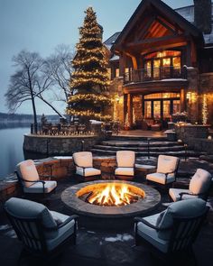 an outdoor fire pit with chairs around it in front of a large house and christmas tree