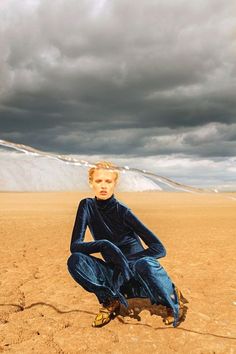 a woman sitting on top of a sandy beach next to a mountain under a cloudy sky