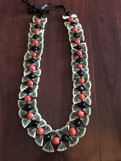 a necklace made out of dollar bills and red beads on a wooden floor with a black ribbon