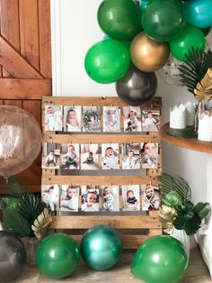 a table topped with balloons and photos next to a wooden crate filled with green, silver and gold balloons