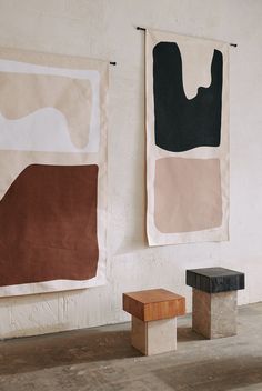 two abstract paintings hang on the wall next to a wooden bench and stool in an empty room