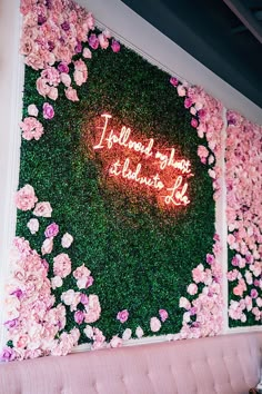 pink flowers and greenery decorate the wall behind a couch with a neon sign that reads, i would make it look like you are in love