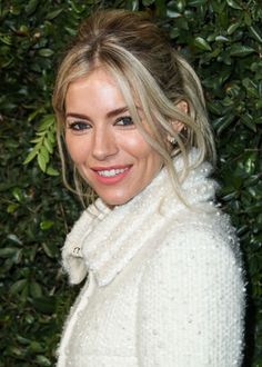 a woman with blonde hair wearing a white coat and smiling at the camera while standing in front of bushes