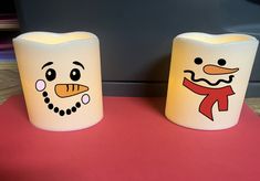 two frosted candles with faces on them sitting on a red tablecloth covered surface