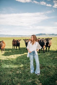 a woman standing in the middle of a field with cows behind her on a sunny day