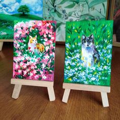 two small paintings on wooden easels sitting on a table next to eachother