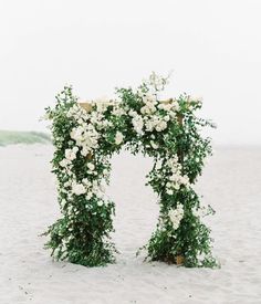 an arch covered in white flowers and greenery on the beach with sand behind it