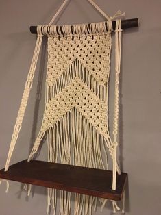 a macrame hanging on a wall with a wooden shelf underneath it and some white yarn