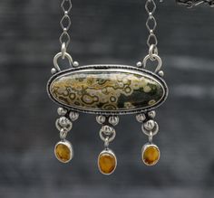 This pendant features a beautiful old stock ocean jasper. It has well defined orbs in a creamy beige with pops of yellow and a green base color. I have set it in my double bezel pendant that I like to do with three dangles of this beautiful mandarine citrine which is the perfect accent to the yellow rings in the orbs in the jasper. It pulls the color out so nicely. I have added a complimentary pattern to the back of the piece that mimics the stone. Top of the bail to bottom of the pendant it is Luxury Artisan Necklace With Patina, Bezel Pendant Ideas, Ocean Jasper Gemstone Pendant Necklace, Shiny Trinkets, Iolite Necklace, Pmc Jewelry, Jewelry Closet, Silversmithing Jewelry, Bezel Set Necklace