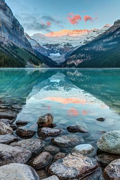 a lake surrounded by mountains with rocks in the foreground and snow capped peaks in the background